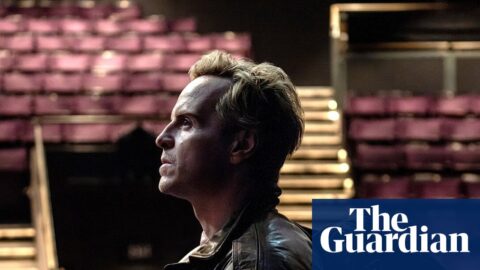 Andrew Scott, in a leather jacket, stands in profile looking upward inside an empty theatre