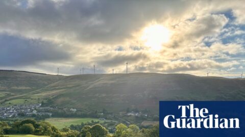 A view across a valley towards sunlit hills with wind turbines and a village off to the left