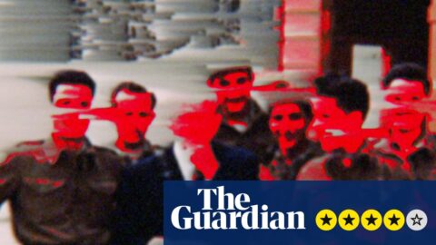Film still showing a group of men posing outdoors, one in a suit holding a hat, red colouring is daubed over their faces